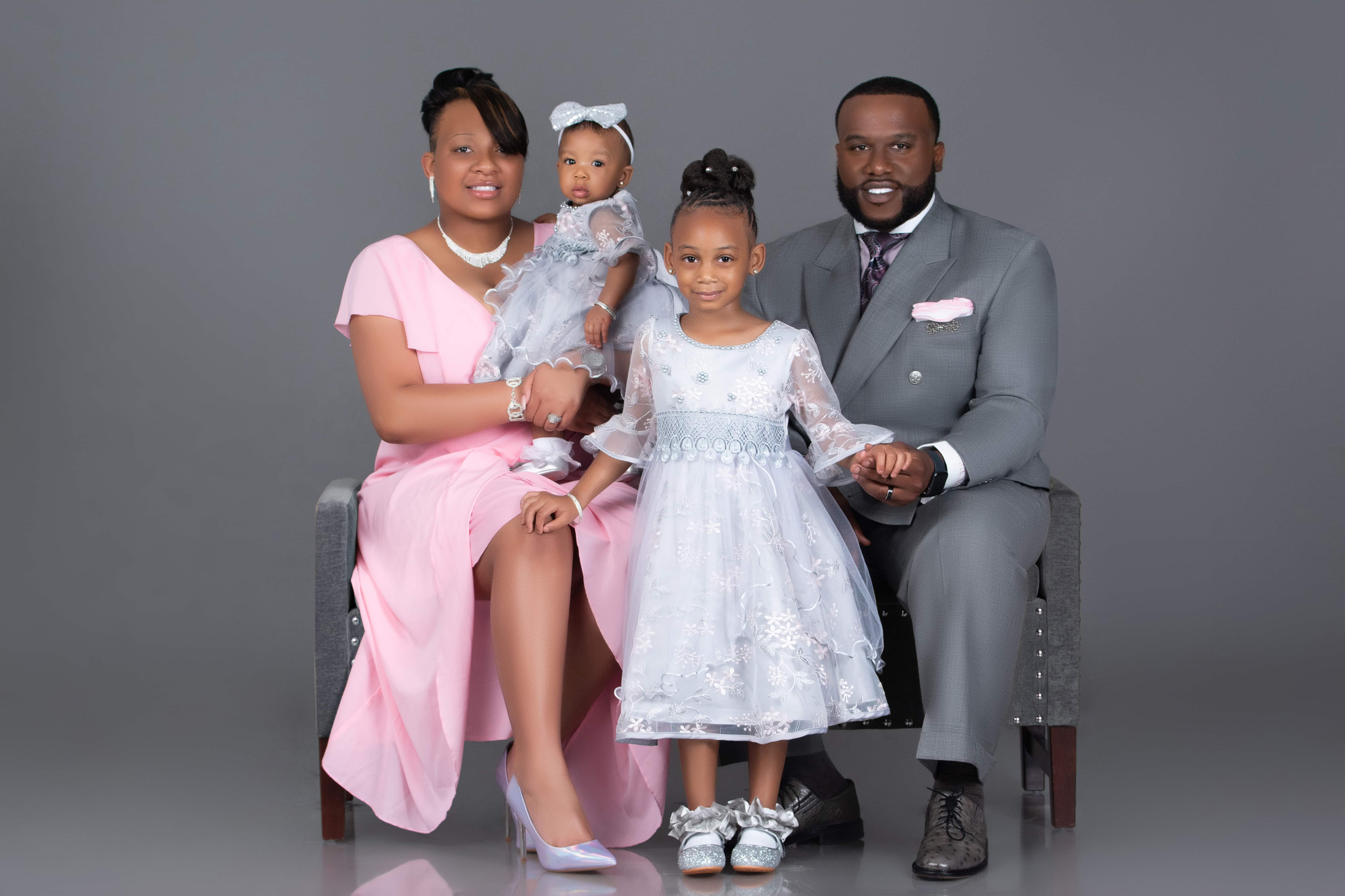 Family with a baby and young daughter in formal pastel outfits posed on a studio bench in Biloxi, MS.