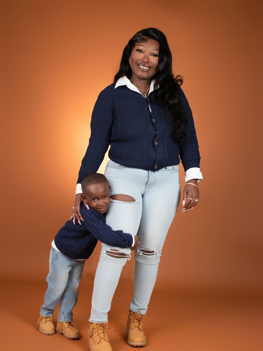 Young son hugging his mother's leg in matching navy sweaters against an orange backdrop in Biloxi, MS.
